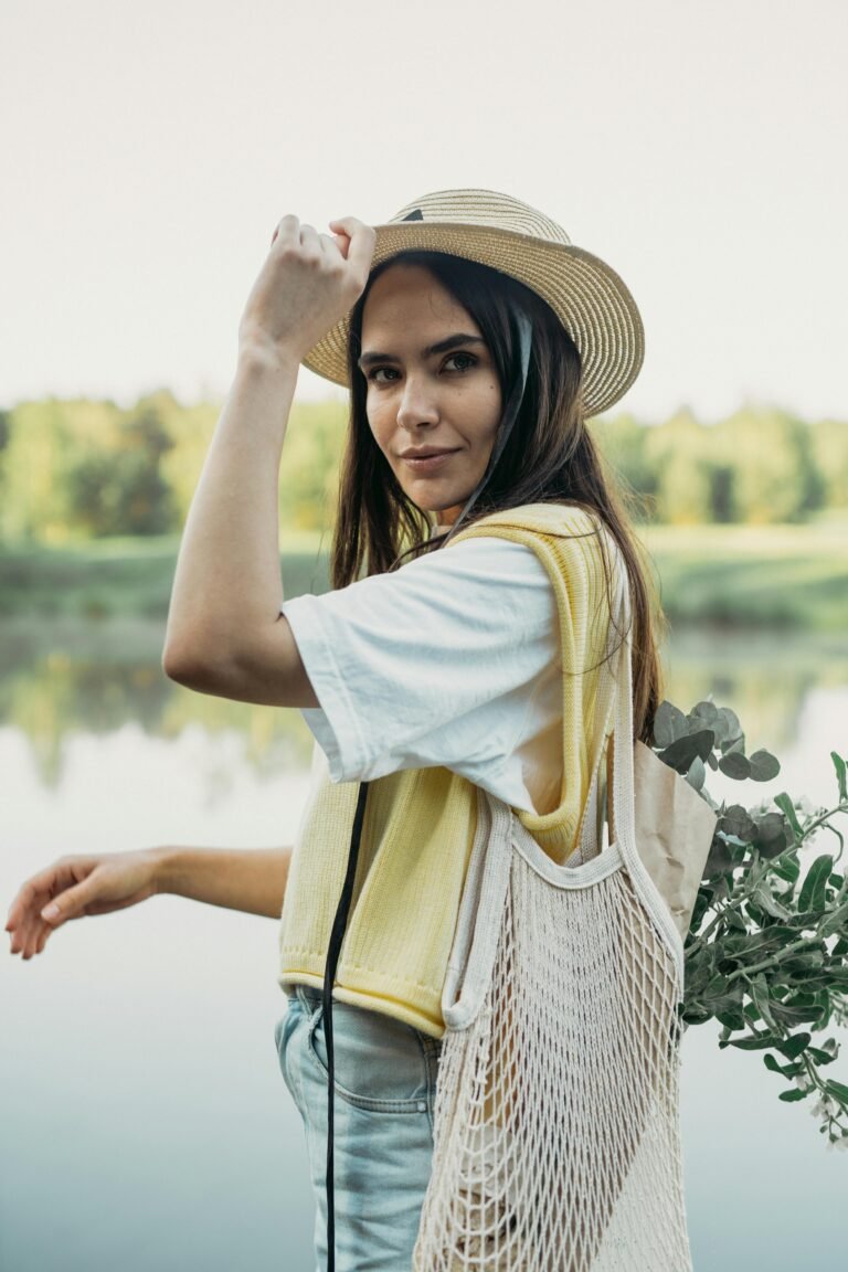 Stylish woman in a straw hat with mesh bag enjoys a peaceful moment by a scenic lake.